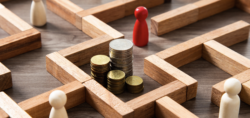 Wooden maze with game pieces trying to reach a stack of coins, symbolizing strategic financial planning and the path to business success